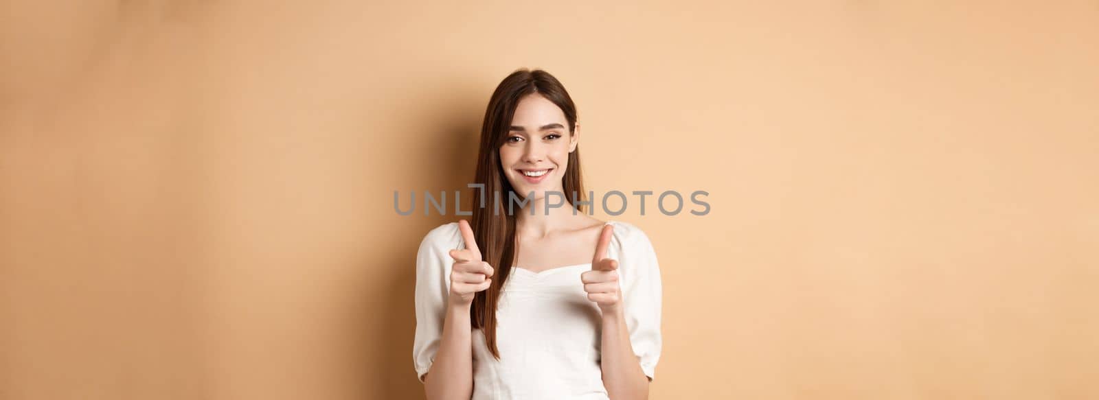 We need you. Smiling young woman pointing fingers at camera to beckon or invite people, standing on beige background by Benzoix