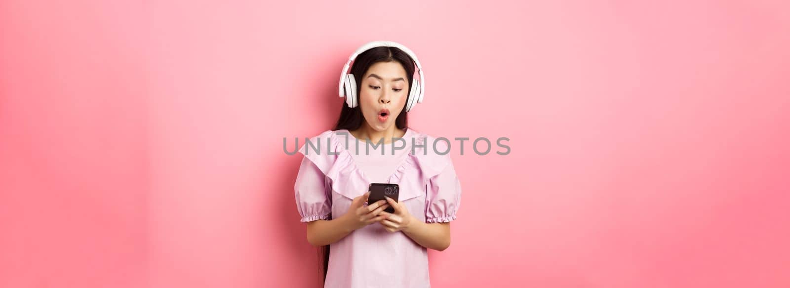 Excited asian girl looking at smartphone screen amused, say wow, listening music in wireless headphones, standing against pink background by Benzoix