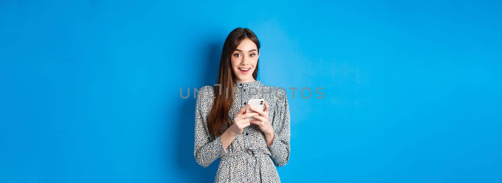 Cute smiling girl with long natural hair, wearing dress, using smartphone and looking happy, standing against blue background by Benzoix