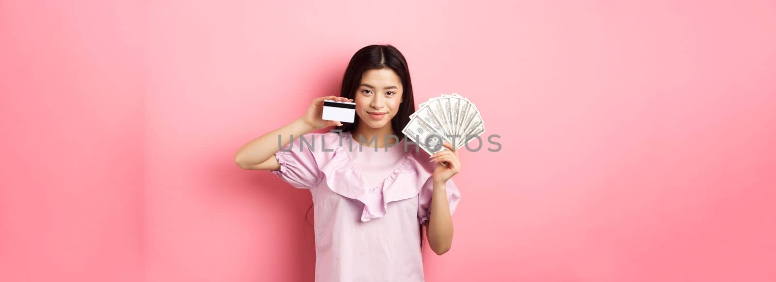 Smiling beautiful asian girl showing money and plastic credit card, giving choice, standing against pink background.