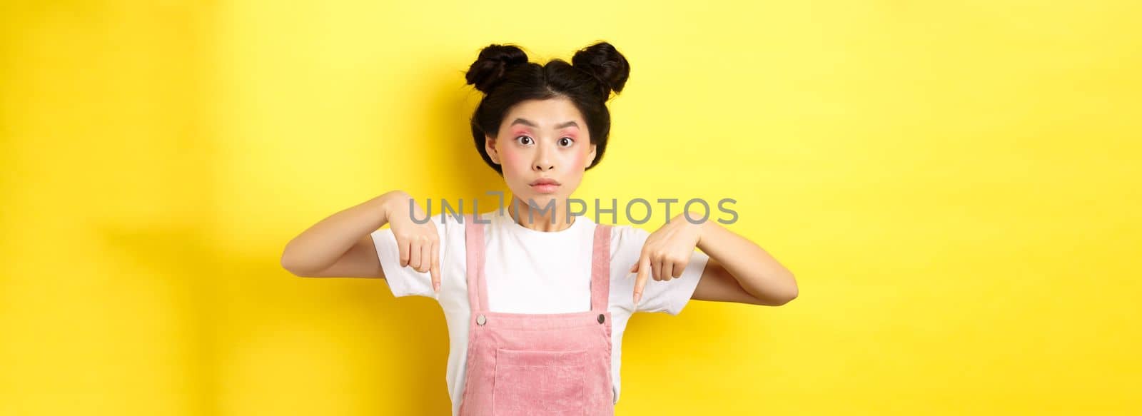Surprised asian woman with beauty makeup, pointing fingers down and looking excited, showing interesting news, yellow background.