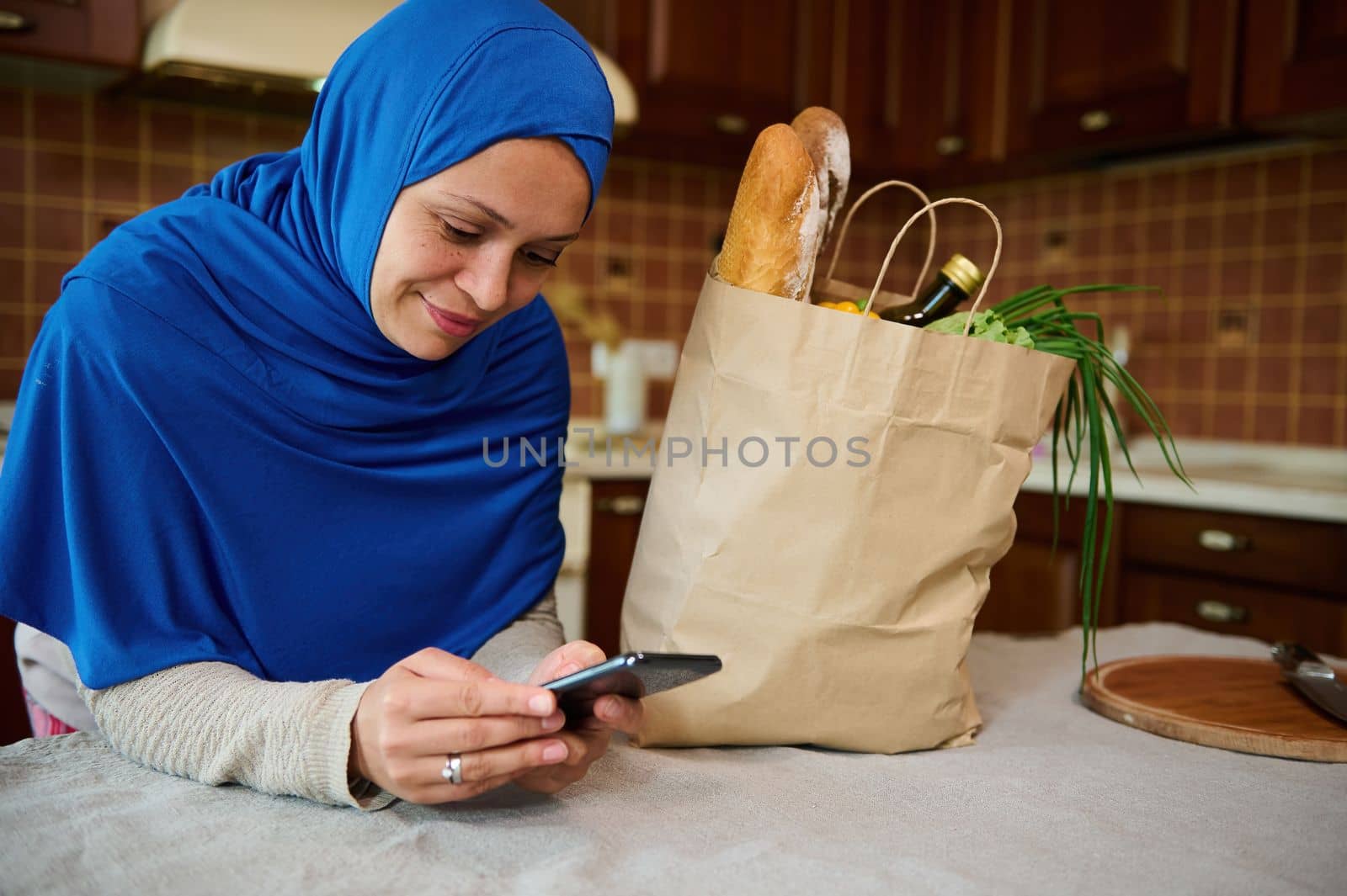 Beautiful Arab Muslim woman in hijab, using smartphone, shopping online, leaning against a kitchen table with a grocery shopping bag with healthy food. Healthy purchasing from grocery. Food delivery.