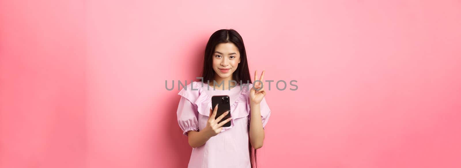 Online shopping. Stylish asian teen girl using smartphone, show peace sign and smiling pleased at camera, standing on pink background.