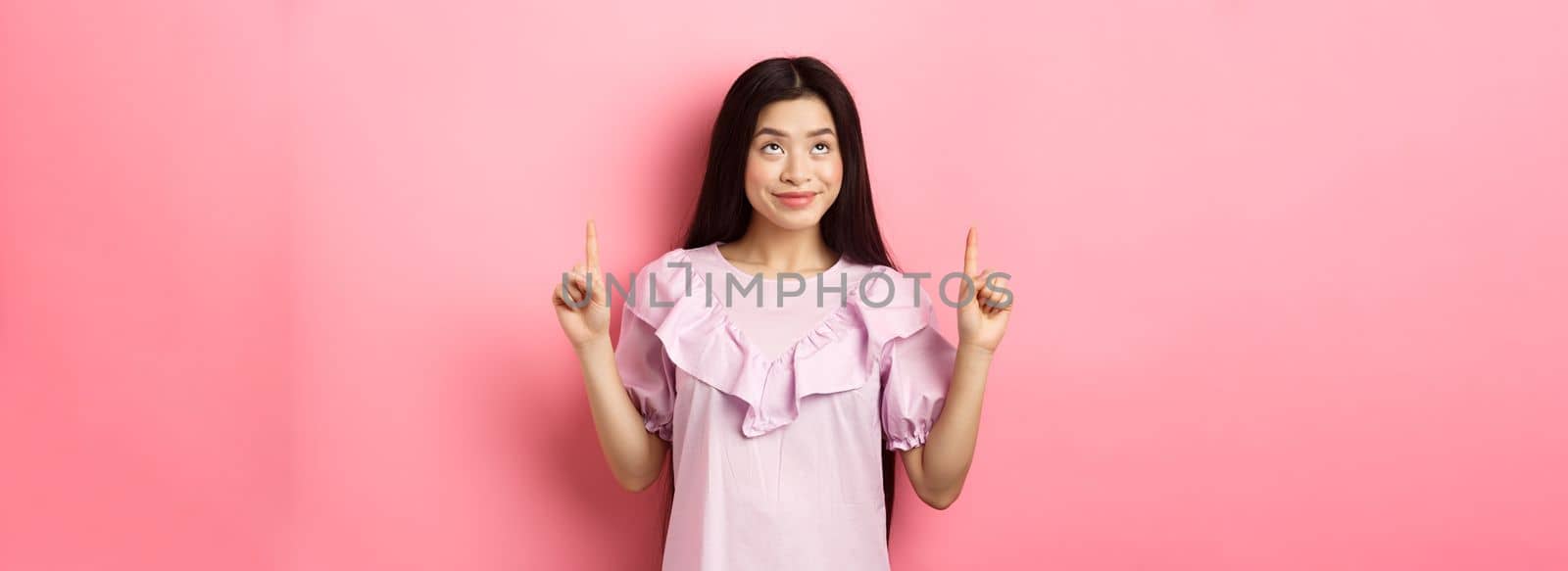 Beautiful smiling asian teen girl pointing fingers up, looking at advertisement with happy face, standing on pink background.