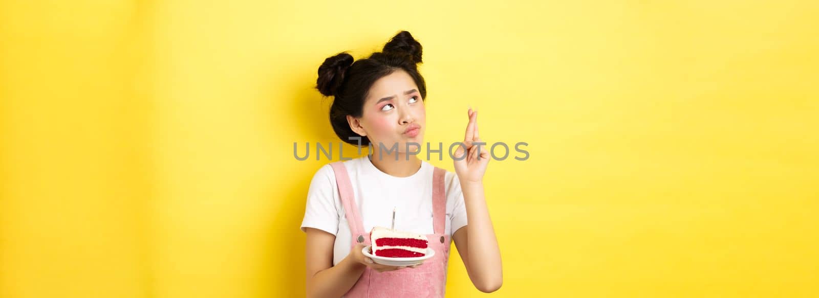 Holidays and celebration. Cute asian birthday girl making wish at party wish cake and candle, cross fingers and looking up, standing on yellow background.