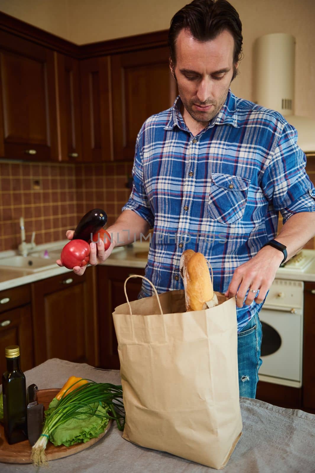 Charming handsome middle-aged Caucasian man lays out purchases in his cozy wooden home kitchen, sorting vegetables and fresh greens while unpacking a grocery shopping bag