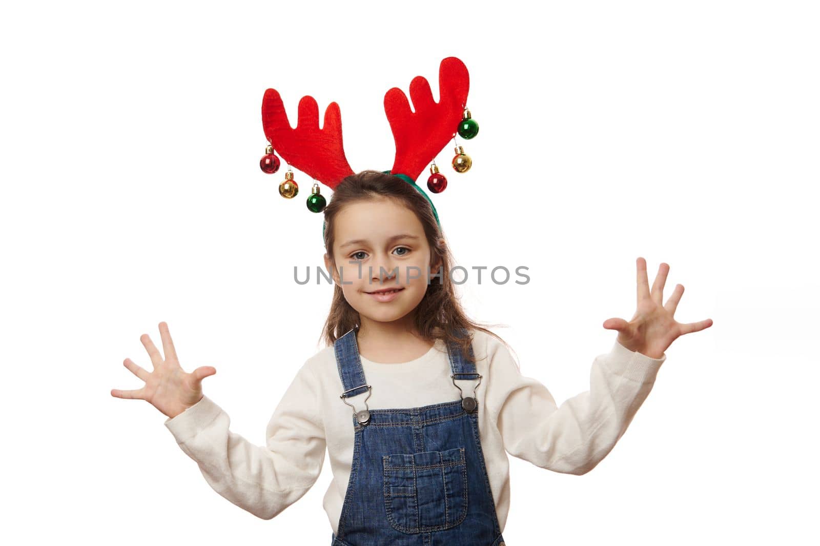 Isolated portrait on white background of a delightful Caucasian baby girl wearing white pull, blue denim overalls and deer antler hoop, expressing joy and happiness, cutely smiling looking at camera