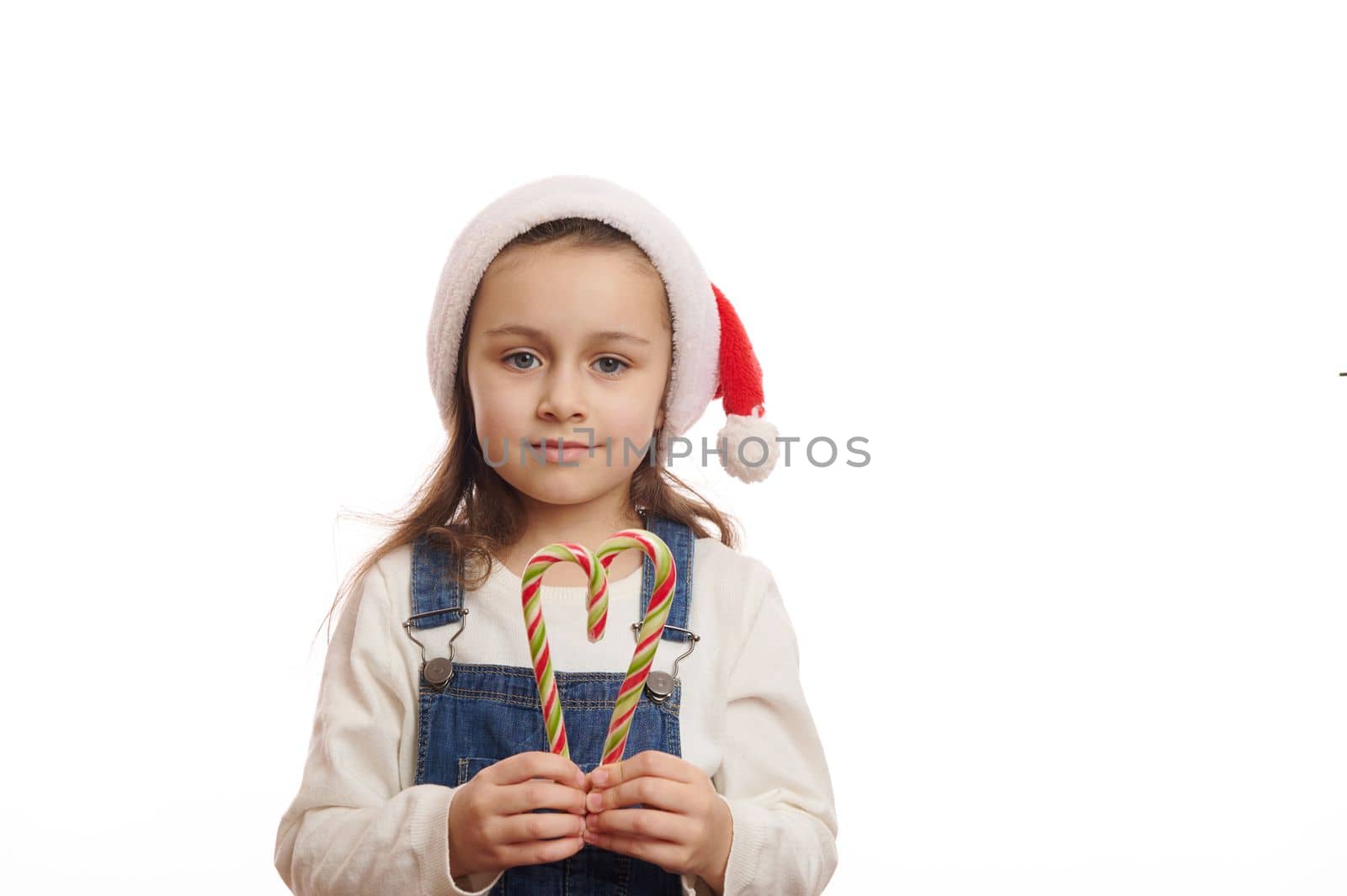 Lovely blue-eyed European little child girl in santa hat, holds striped christmas lollipops in the shape of heart, looking at camera isolated on white background. Copy space for christmas advertising