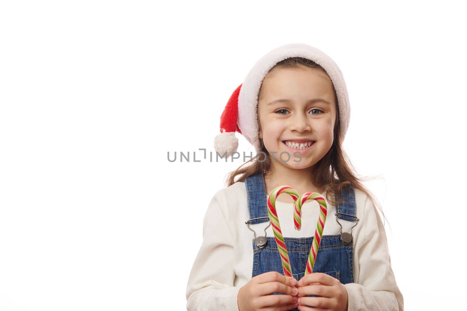 Lovely child, Caucasian baby girl in Santa hat, smiling a cheerful toothy smile at camera, posing with red and green striped candy canes over white background. Merry Christmas and New Year's concept
