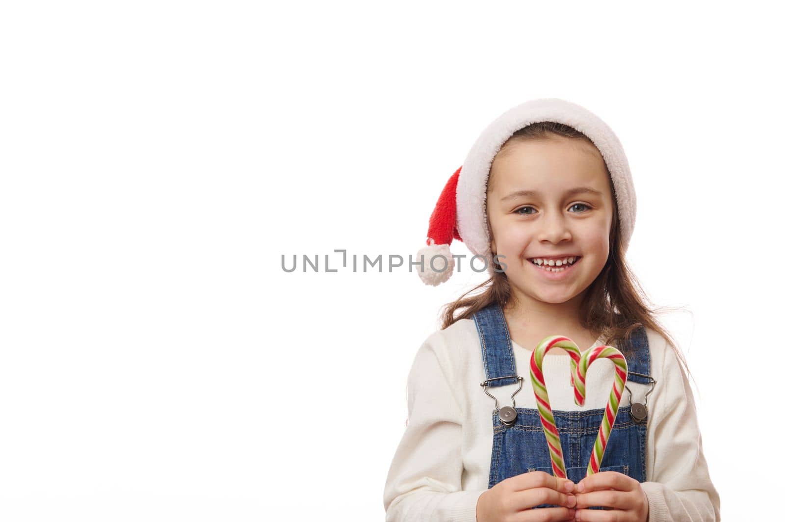 Beautiful blue-eyed Caucasian little girl with long hair, in Santa hat, holds Christmas striped lollipops, sweet candy canes in the shape of heart, smiles a cheerful toothy smile at camera. Copy space