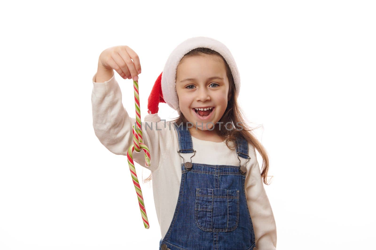 Caucasian smiling cheerful little girl wearing blue denim overalls and red Santa hat, playing with striped red and green candy canes lollipops on white background. Christmas New Year winter Holidays