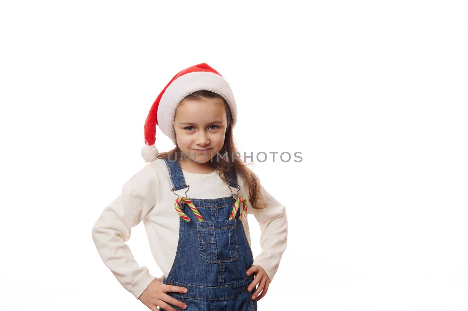 Adorable Caucasian little child girl wearing Santa hat, posing with hands on waist, with Christmas striped candy canes on the pocket of his denim overalls, looking at camera, against white background