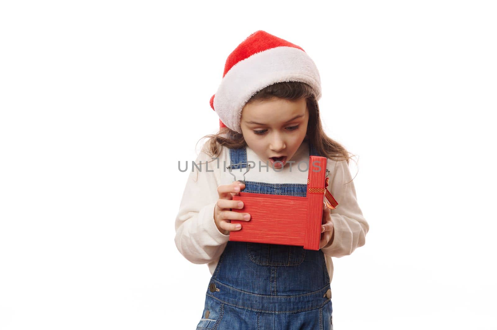 It's time to open Christmas New Year presents. Caucasian little girl in Santa hat, with excited emotions, unpacking a red gift box with cute surprise. Enjoy the magic atmosphere of winter holidays