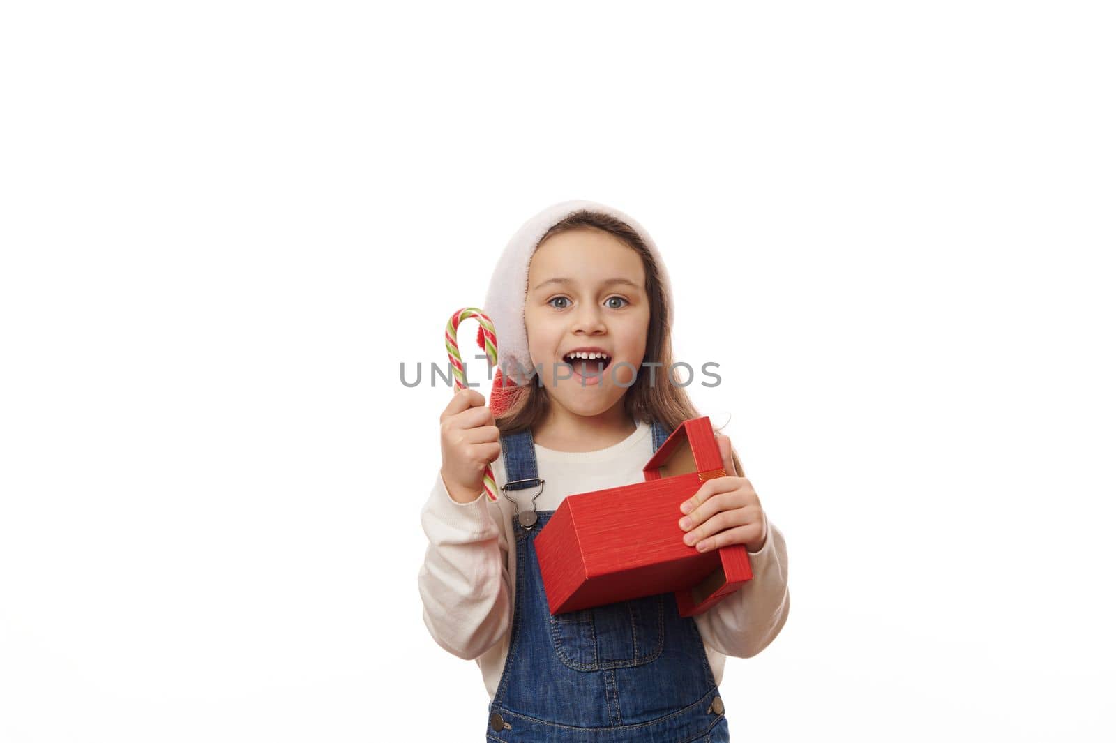 Delightful baby girl in Santa hat, smiling a beautiul toothy smile looking at camera, posing with striped lollipop candy cane and red gift box, on white background. Merry Christmas and Happy New Year