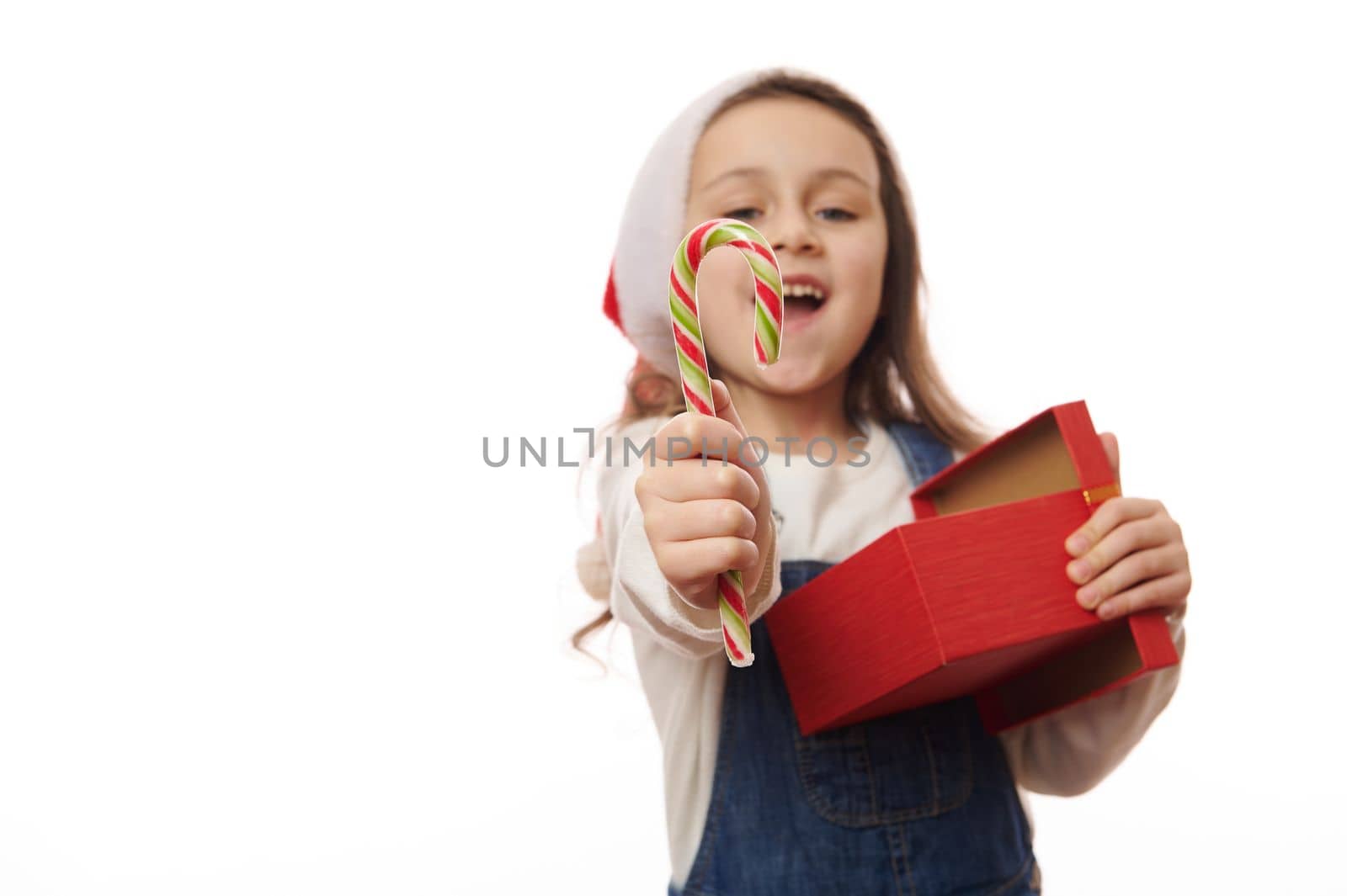 Focus on red green striped lillipop in the hands of a blurred overjoyed happy smiling child girl in Santa hat, with gift box, holding out a sweet candy cane to camera, isolated over white background