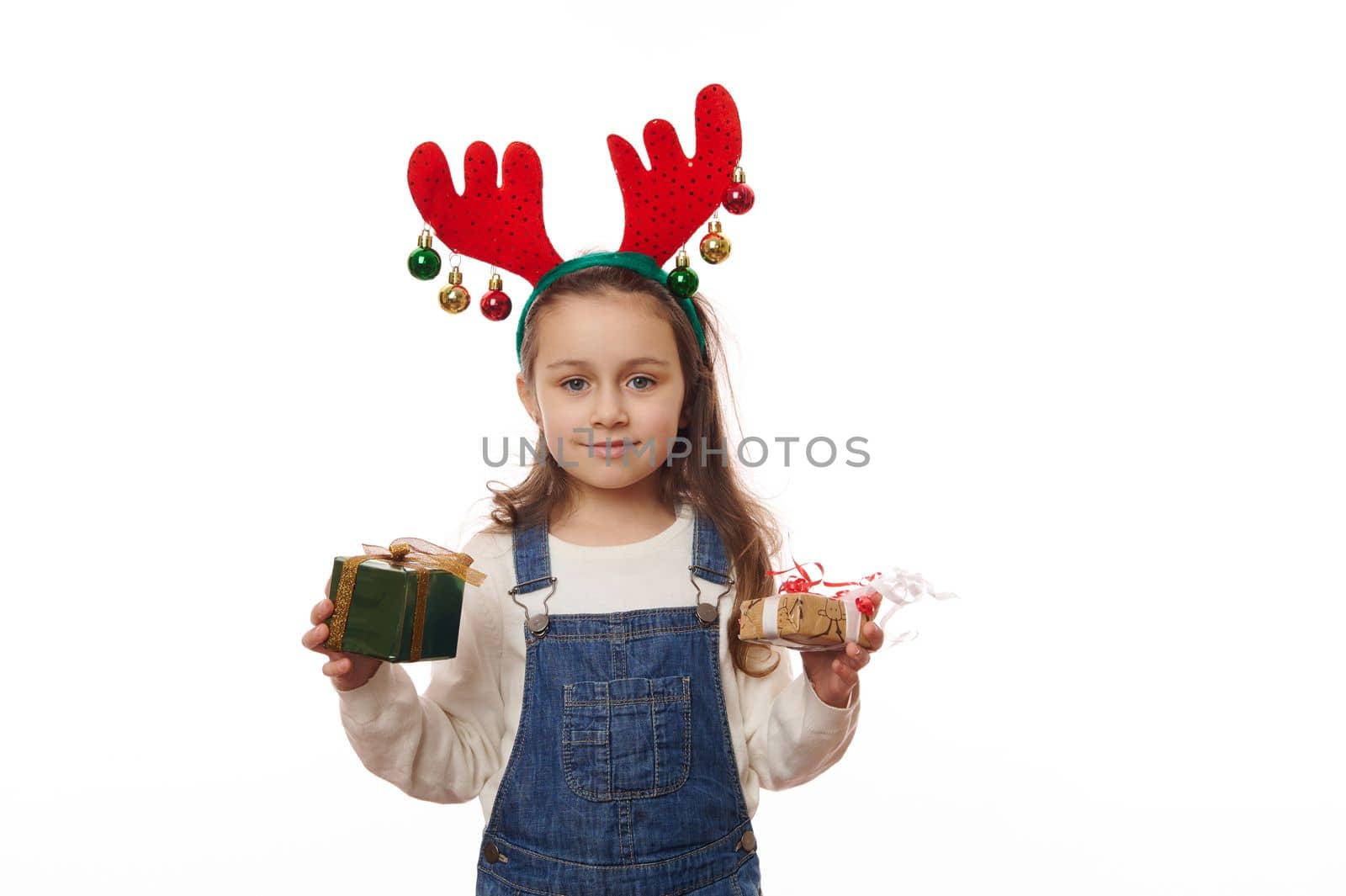 Cheerful Caucasian little girl in deer antler hoop, posing with happy Christmas presents over white background. Happy atmosphere of upcoming winter holidays. Boxing Day. It's time to open Xmas gifts