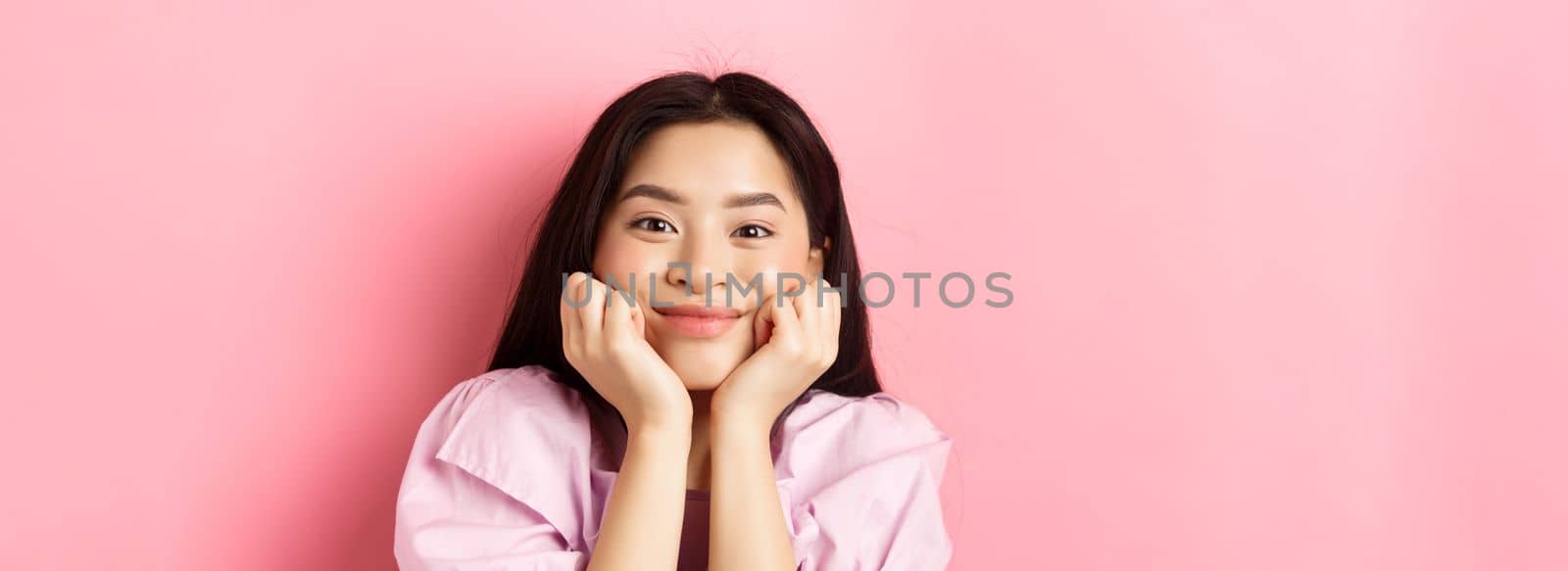 Close-up of excited asian girl listening with interest, smiling amused and looking at camera, standing against pink background by Benzoix