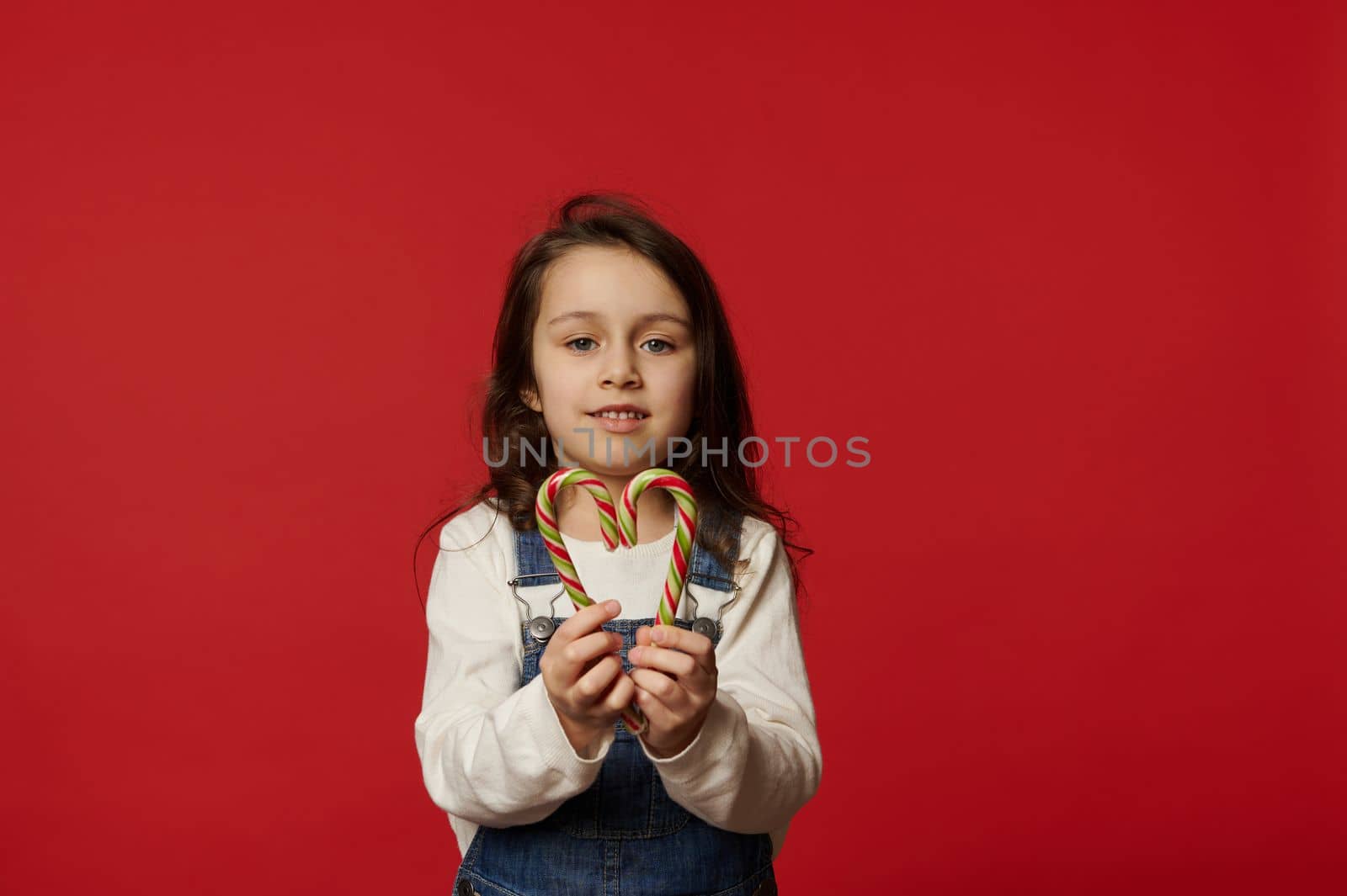 Beautiful Caucasian blue-eyed little child girl with long hair, wearing white pull and blue denim overalls, holding striped candy cane, Xmas lollipops in the shape of heart, isolated on red background
