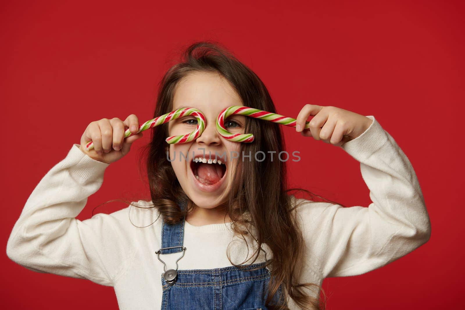 Funny amazed cheery little child girl, looking through striped lollipops like eyeglasses, laughing looking at camera, isolated over red background with free advertising space for text. Merry Christmas