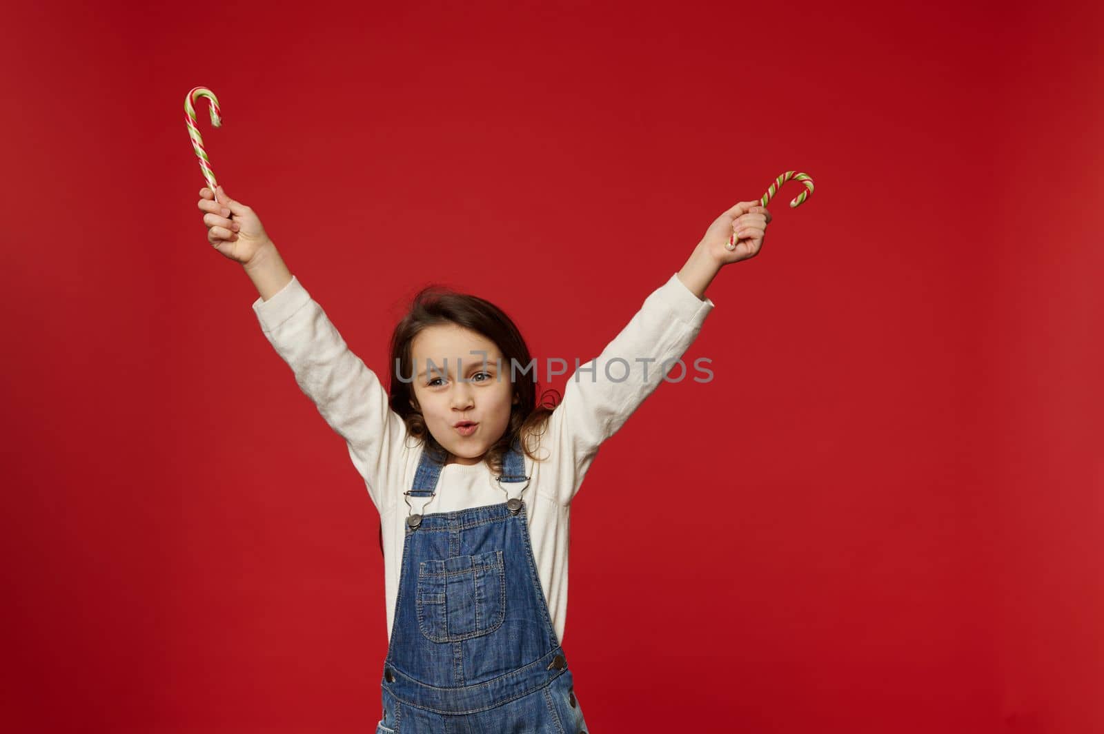 Cherry baby girl in white pullover and blue overalls holding Christmas candy canes in her outstretched hands, smiling enjoying the magical festive atmosphere of upcoming winter events, red background