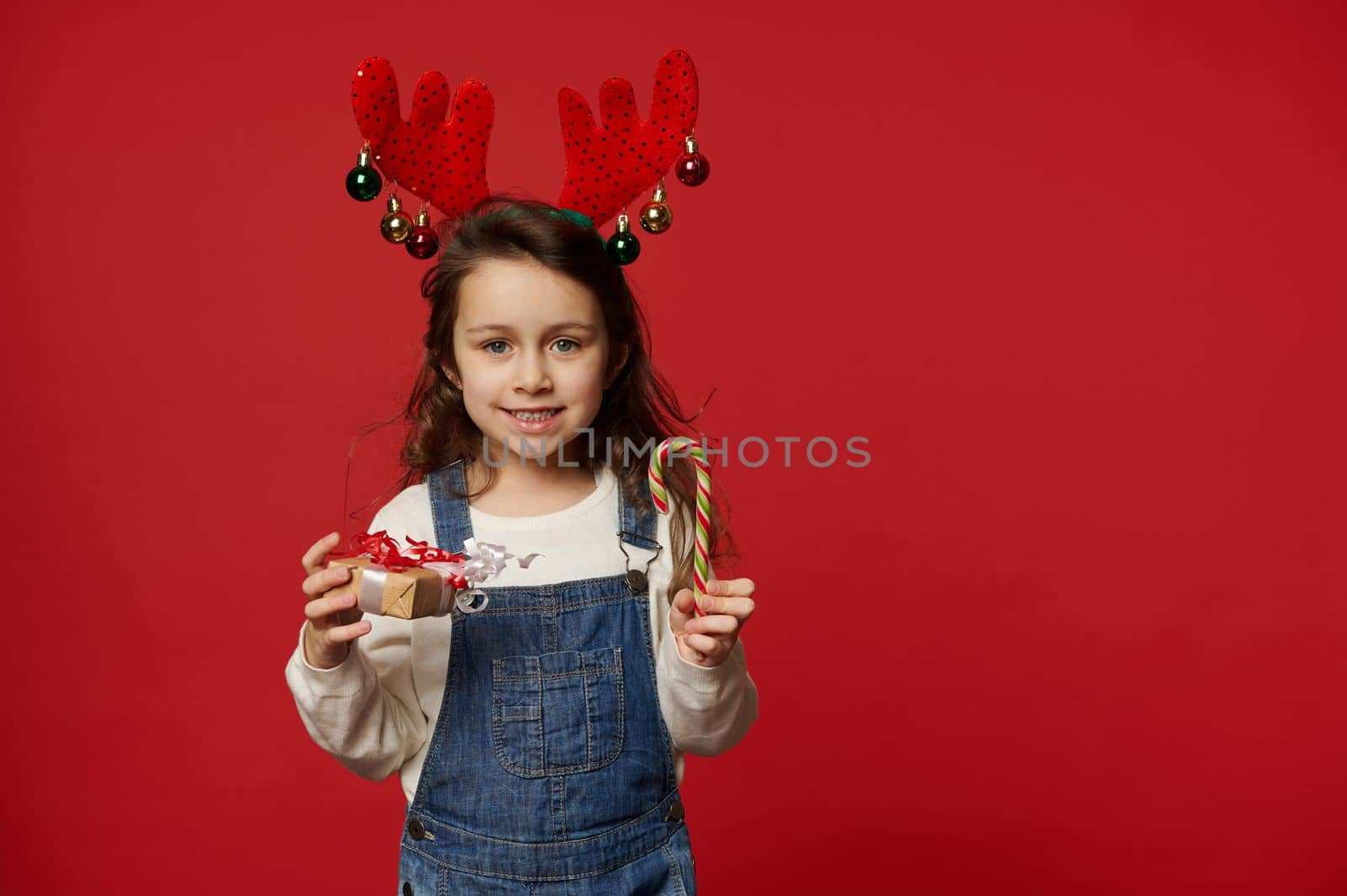 Adorable child, lovely little girl with long hair, wearing deer antler hoop and blue denim overalls, smiles looking at camera, posing with candy cane and Christmas present on red background Copy space