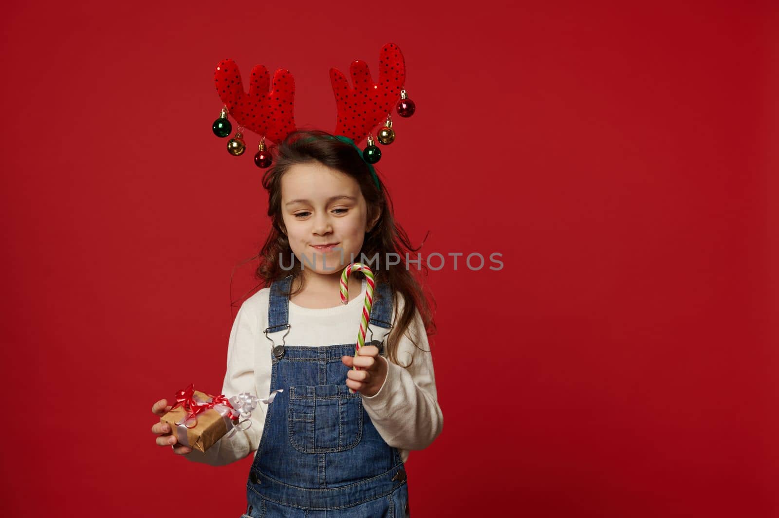 Caucasian cheerful lovely little girl, wearing deer antler hoop, white pullover and blue denim overalls, posing with stripped red and green lollipop - candy cane and Christmas present, red background