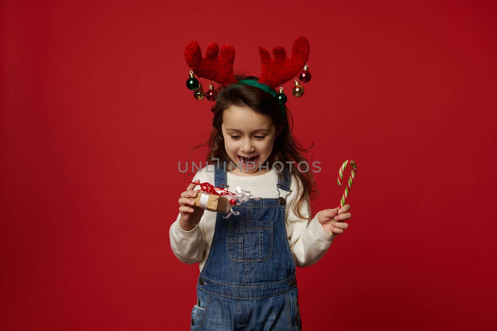 Adorable child little girl, wearing deer antler hoop, posing with stripped red and green lollipop - candy cane and Christmas present, rejoicing at upcoming winter holidays, isolated on red background
