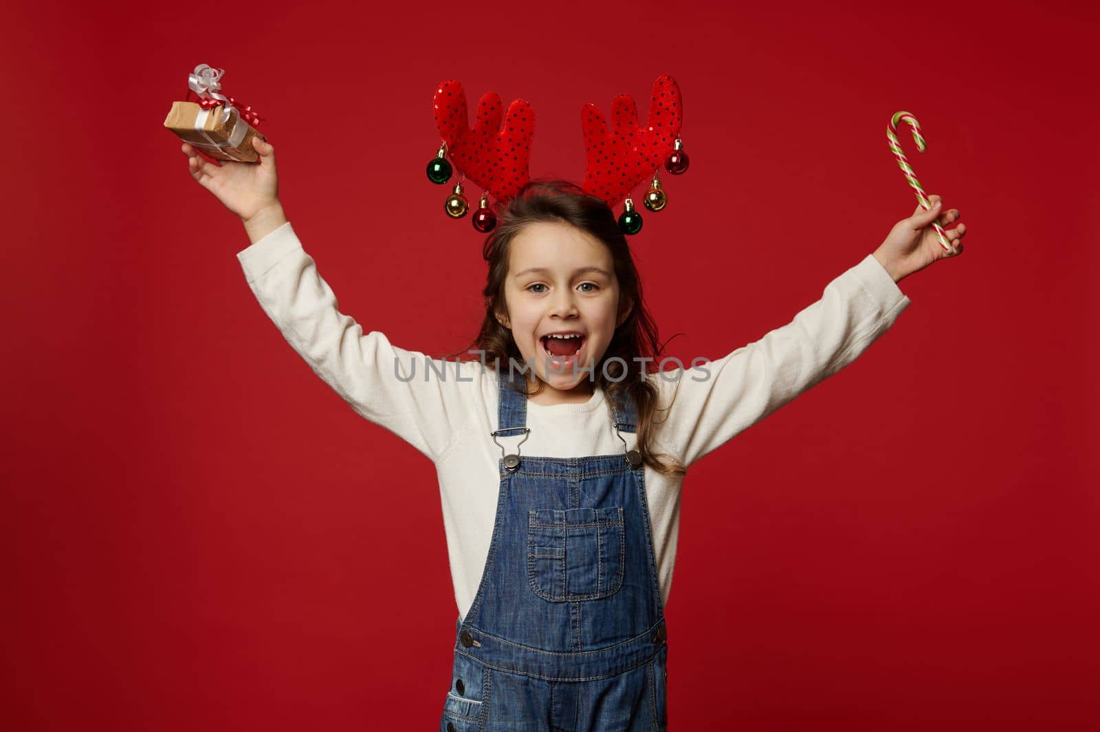 Overjoyed kid, lovely little girl in deer antler hoop, raising her arms up, holding Christmas gift and candy cane, isolated over red background with copy space, enjoying the upcoming winter holidays