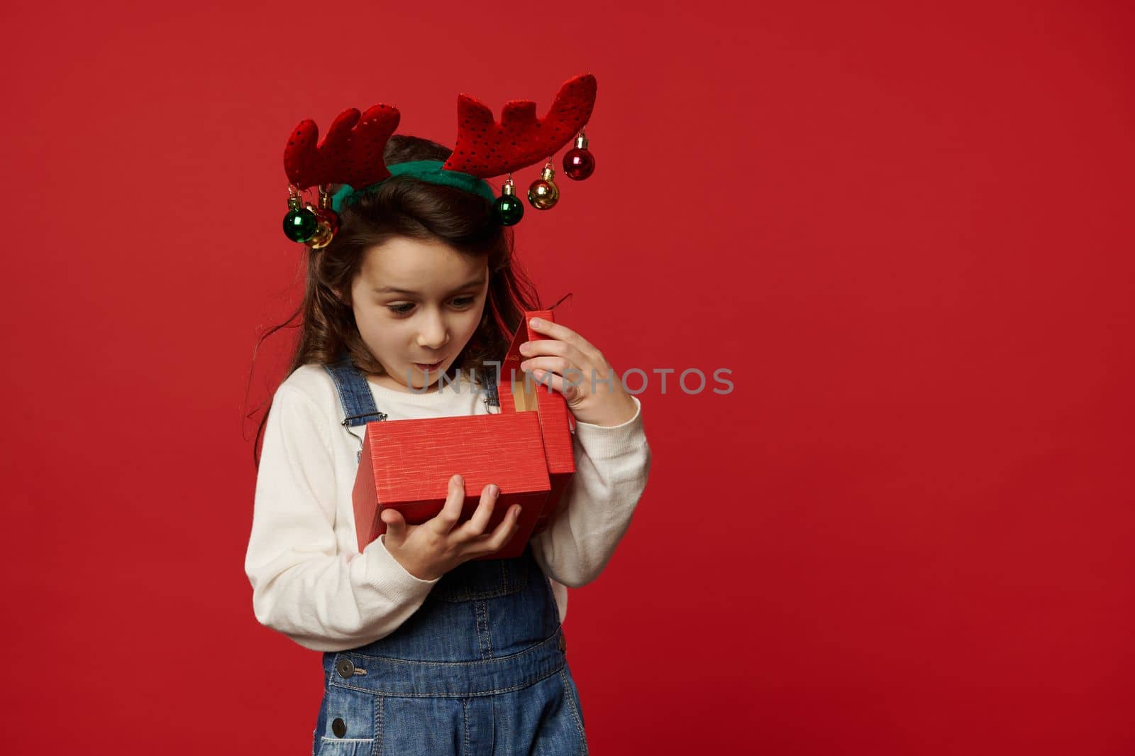 Surprised Caucasian baby girl wearing deer antler hoop, expressing happiness and excitement while unpacking red gift box with happy present for Christmas or New Year, over colored backdrop. Boxing Day