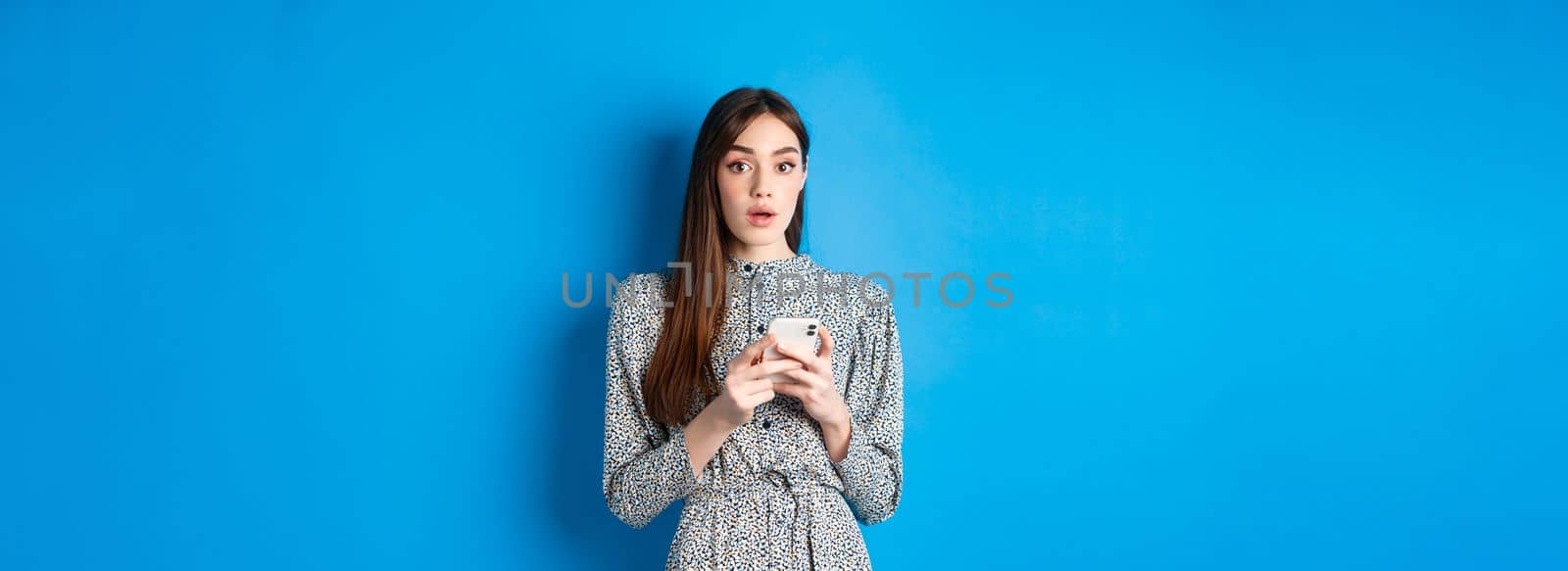 Woman look surprised after using mobile phone, standing on blue background.