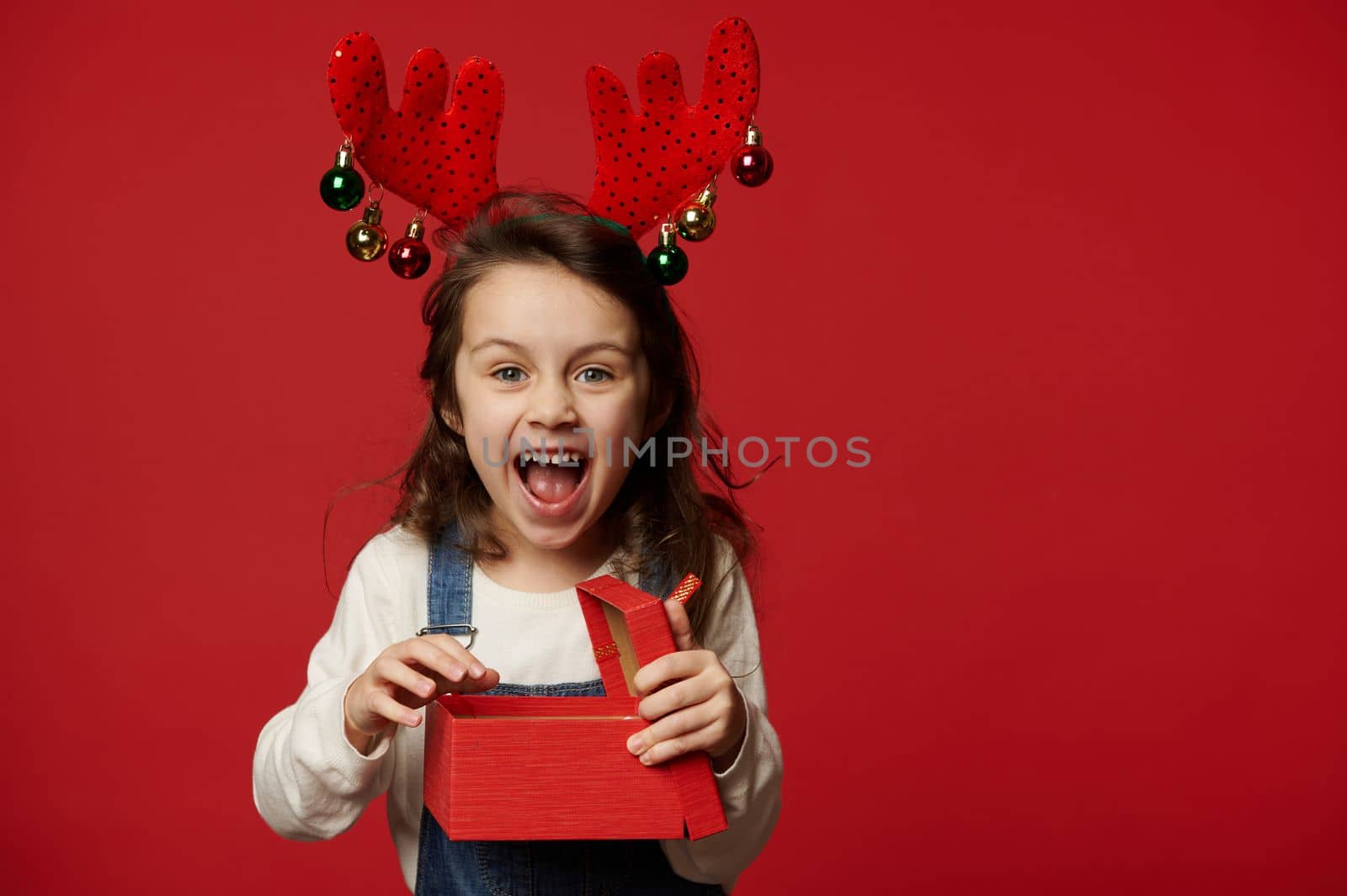 Overjoyed mischievous baby girl wearing denim overalls, white pullover and deer antler hoop, laughing looking at camera, expressing positive emotions, posing with Christmas present on red background
