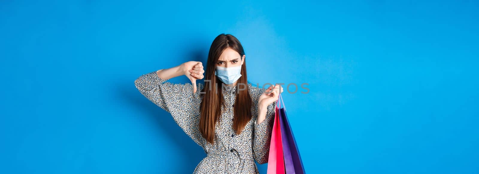 Covid-19, pandemic and lifestyle concept. Angry client in medical mask showing thumb down in dislike, frowning upset, holding shopping bags, blue background.