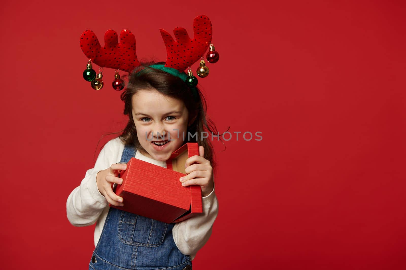 Mischievous funny little child girl in deer antler hoop, white pullover and blue denim overalls, smiling looking at camera, unpacking gift box with surprise for Christmas, on red background. Ad space