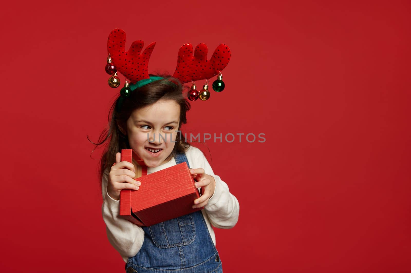 Mischievous cheery baby girl, wearing deer antler hoop, smiling looking aside at copy advertising space, unpacking Christmas or New Year gif box with presents, on red background. Happy winter holidays