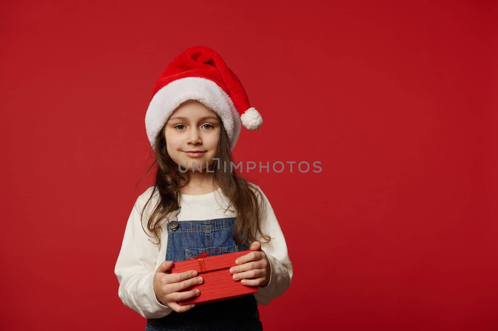Lovely Caucasian little child girl in Santa hat, white pullover and blue denim overalls, smiling looking at camera, posing with cute gift box with surprise for Christmas, on red background. Ad space