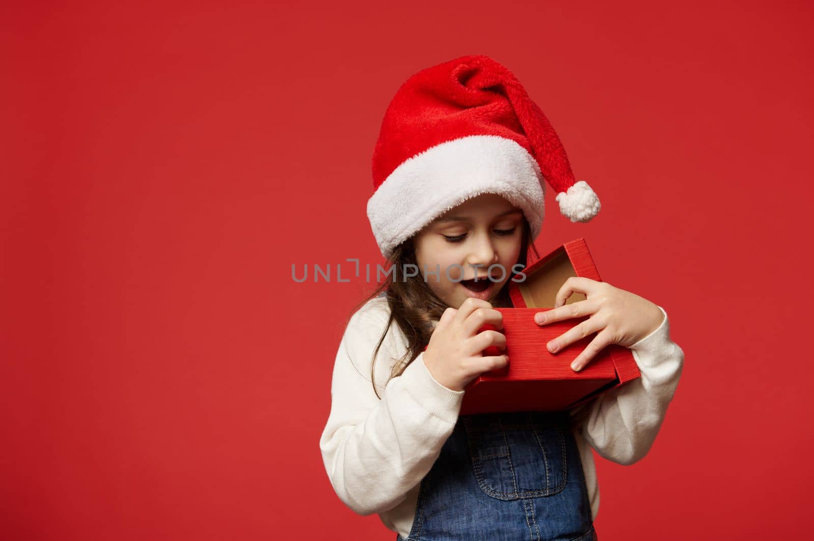 Delightful Caucasian 5 years old little child girl in Santa hat, expressing surprise and positive emotions while unpacking Christmas present over red background. Boxing Day. Happy New Year concept
