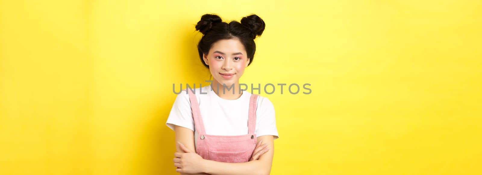 Beauty asian girl with glamour makeup, cross arms on chest and smiling tenderly at camera, standing on yellow background by Benzoix