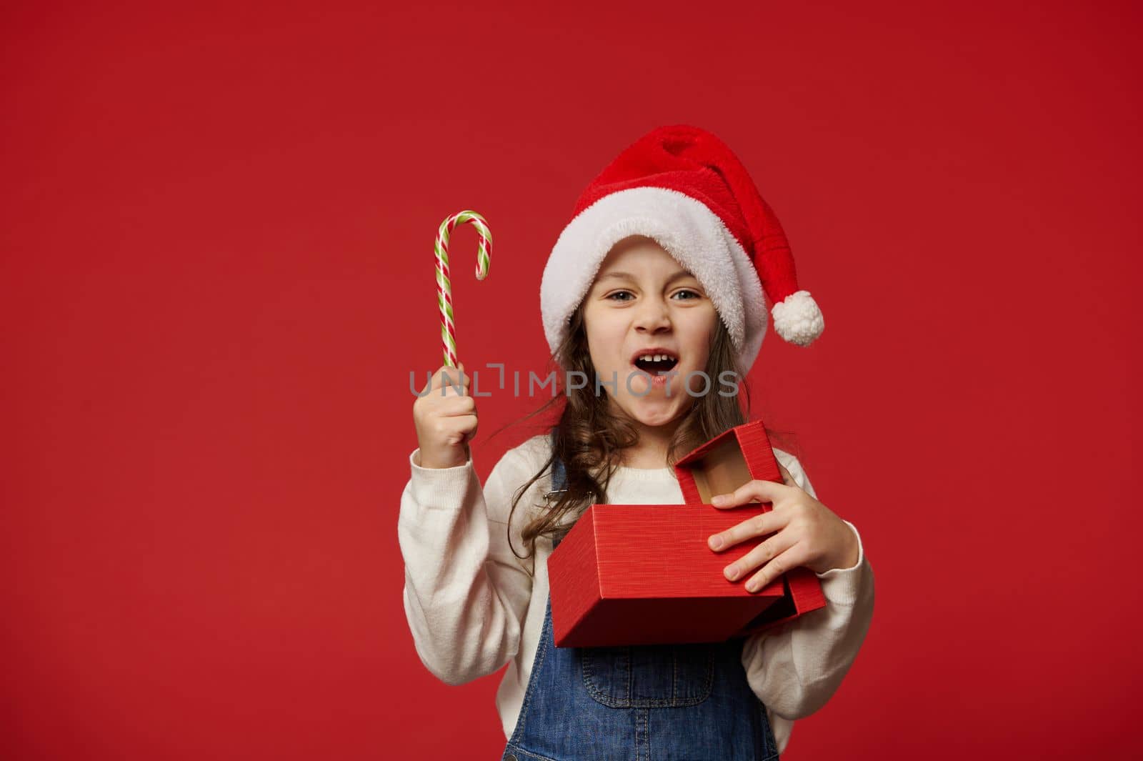 Surprised little girl wearing Santa hat, holding candy cane and red gift box with happy present for Christmas, expressing happiness and joy at upcoming winter holidays, red background. Merry Christmas