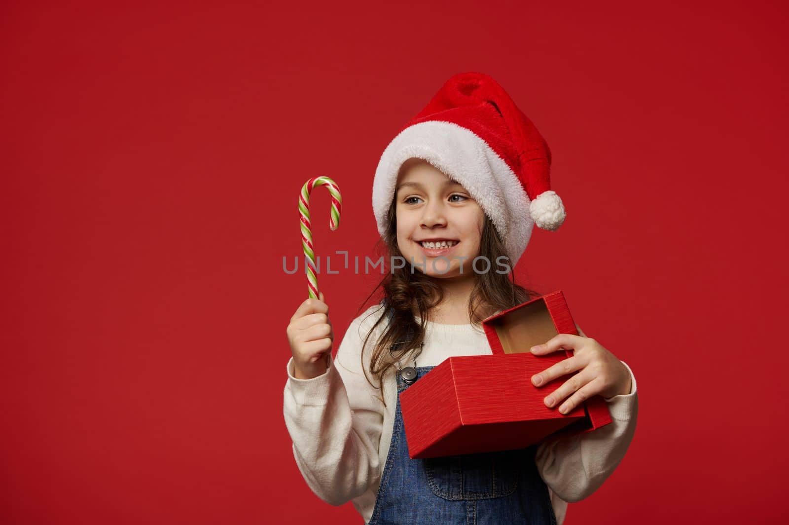 Caucasian happy 5 years old child, lovely little girl in Santa hat, smiling toothy smile looking at candy cane, posing with Christmas present on red background. Copy ad space. Winter holidays New Year