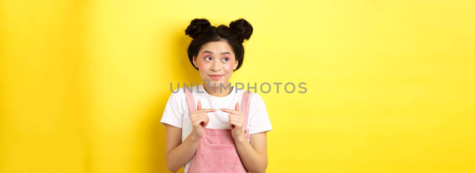 Summer lifestyle concept. Cute shy asian girl looking away and smiling silly, avoiding eye contact, standing against yellow background.