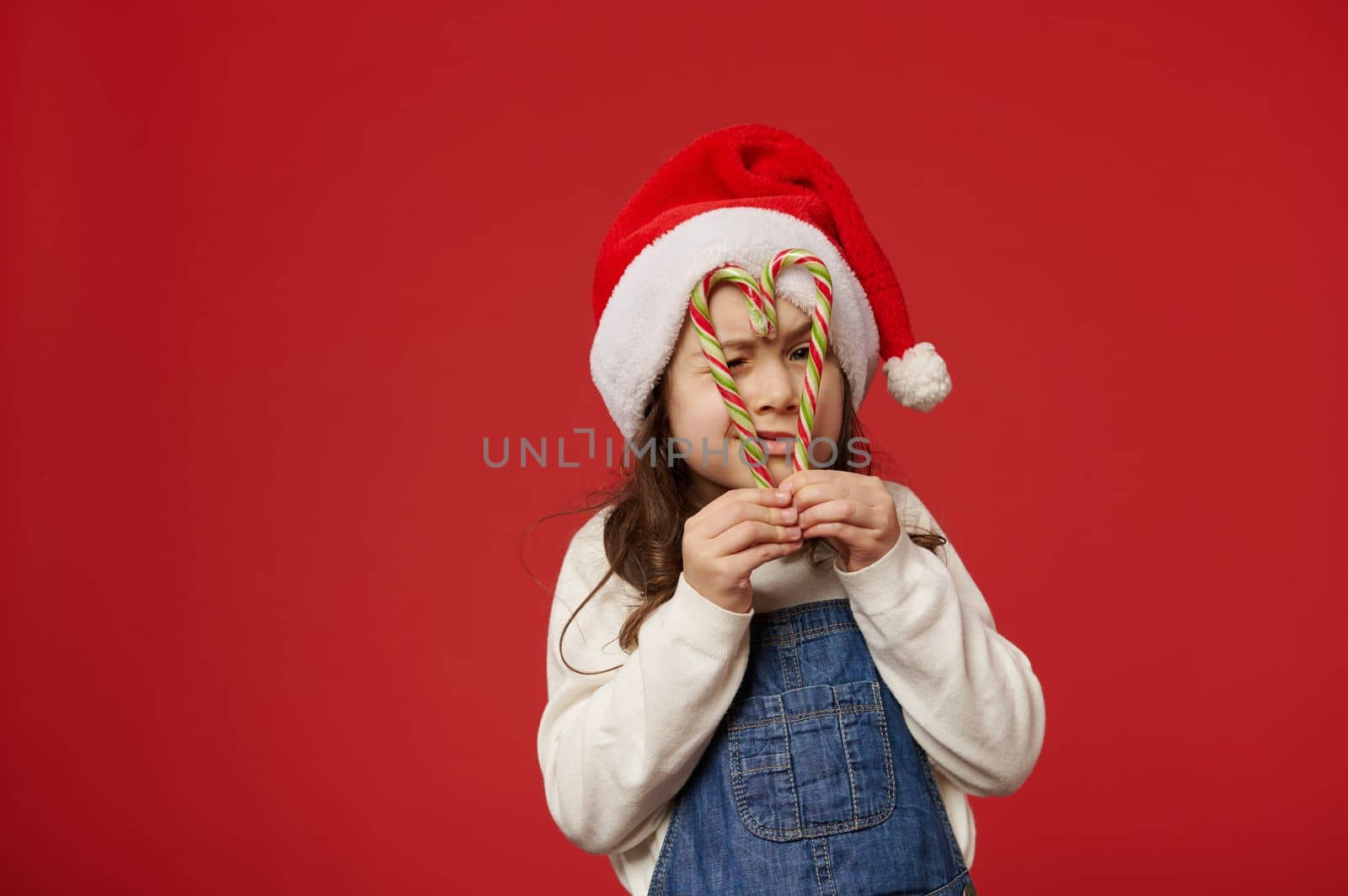 Adorable little child girl in Santa Claud hat, looking at camera through striped lollipops like eyeglasses, isolated over red background with free advertising space for text. Merry Christmas