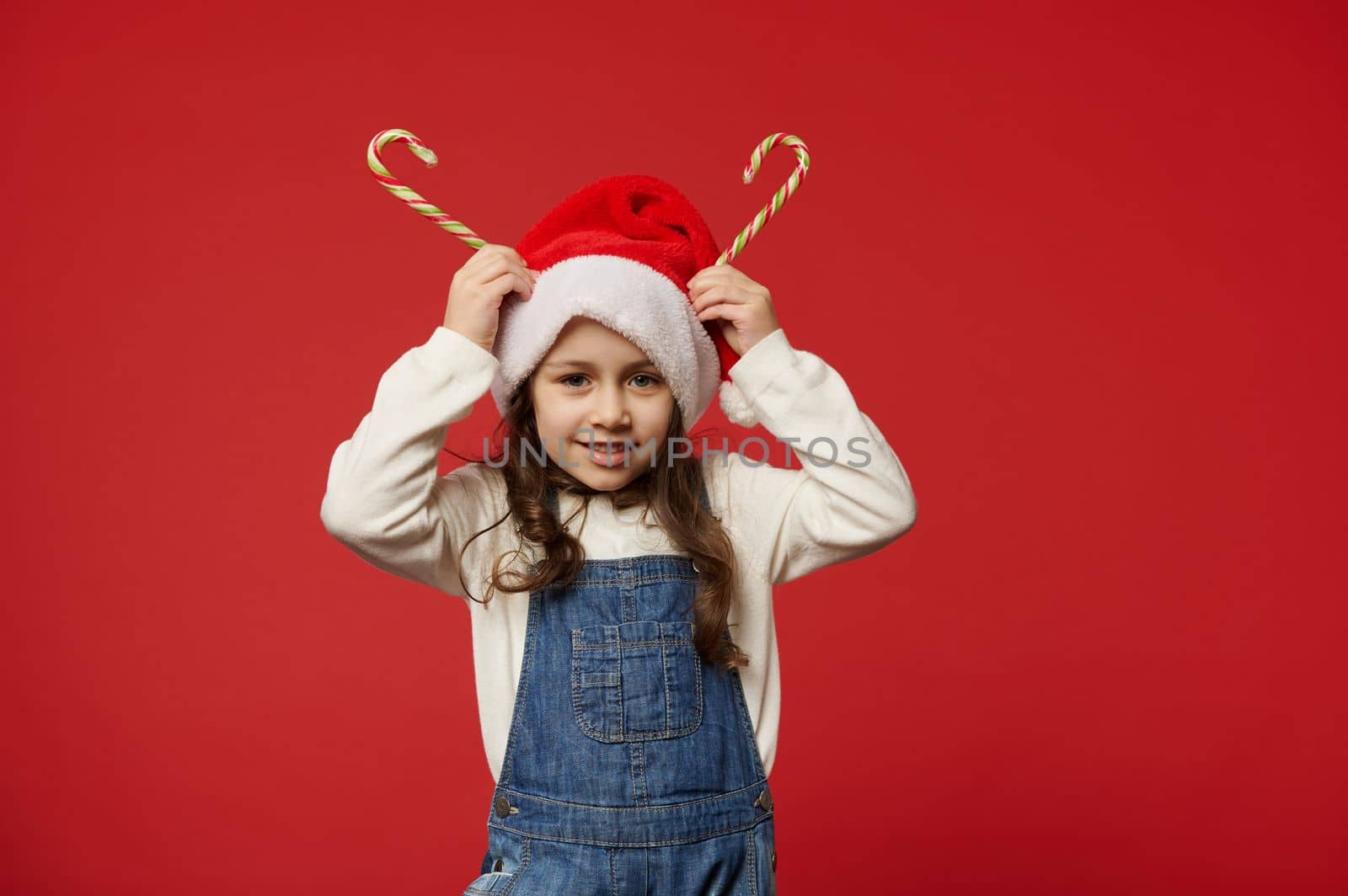 Joyful little girl with beautiful long hair, wearing white pullover, blue denim overalls and Santa Claus hat, holding striped lollipops on her head like horns, smiles looking at camera, red background