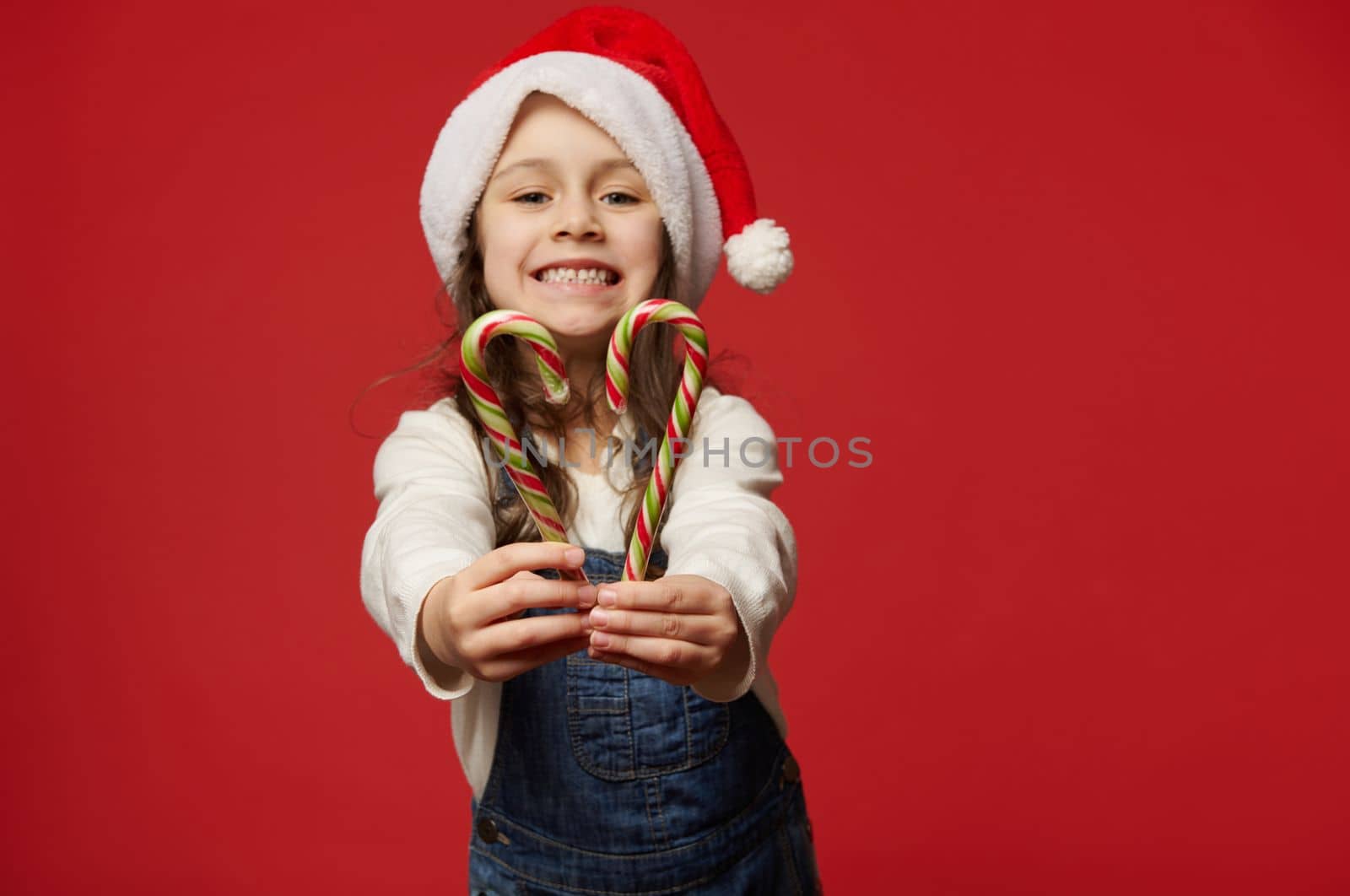 Details: hands of a blurred little child girl in Santa hat, smiling a beautiful cheerful toothy smile, holding out at camera red green stripped lollipops, sweet Xmas candy canes over red background