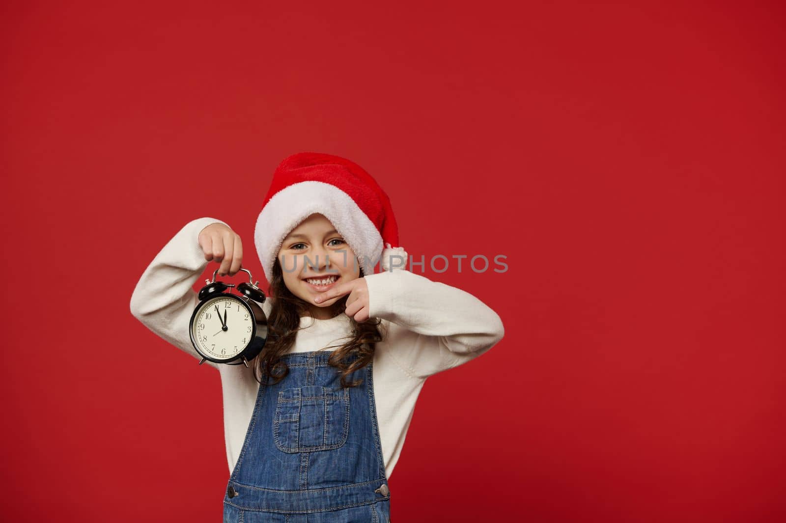 Cheerful little girl in Santa Claus hat, pointing at black alarm clock showing five to midnight, excited by the upcoming Christmas night or New Year celebration, smiling at camera, isolated on red