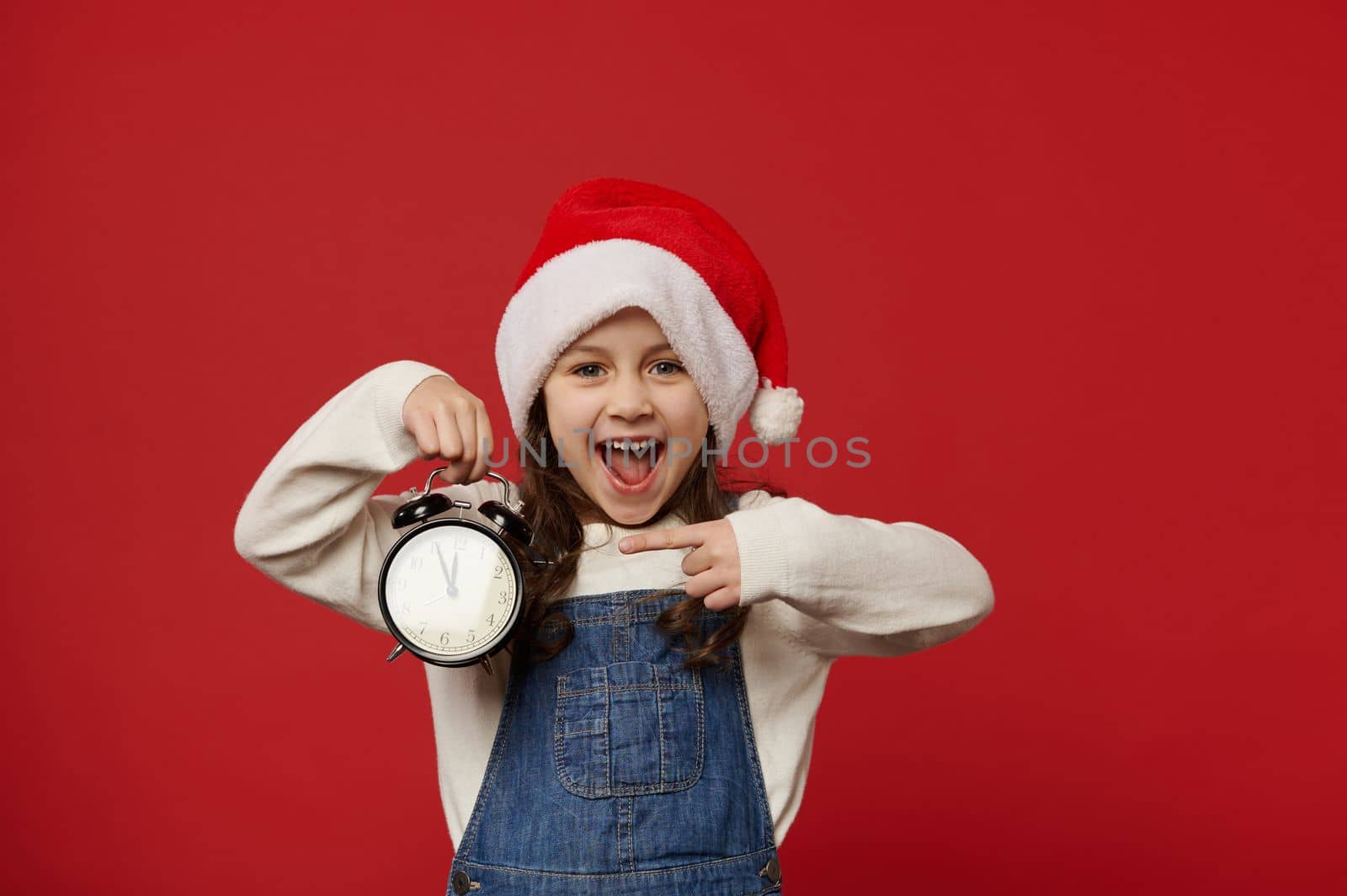 Overjoyed mischievous little girl in Santa hat, pointing at a vintage alarm clock with five to midnight, excited by the upcoming New Year event, isolated over red background. Copy advertising space
