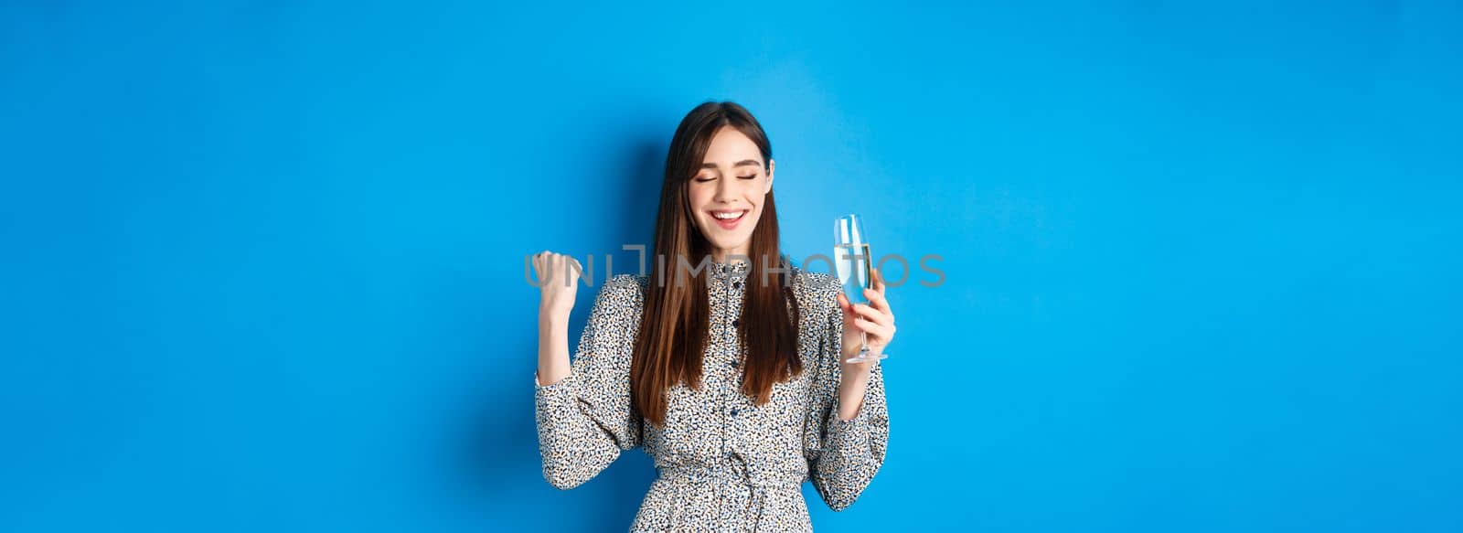 Happy woman celebrating win with glass of champagne, dancing satisfiied, triumphing of victory, standing against blue background.