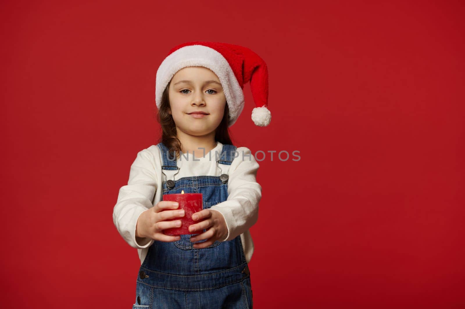 Caucasian lovely child, adorable blue-eyed little girl wearing Santa hat and blue denim overalls, cutely smiling looking at camera, holding out a lit Christmas candle, isolated on red background
