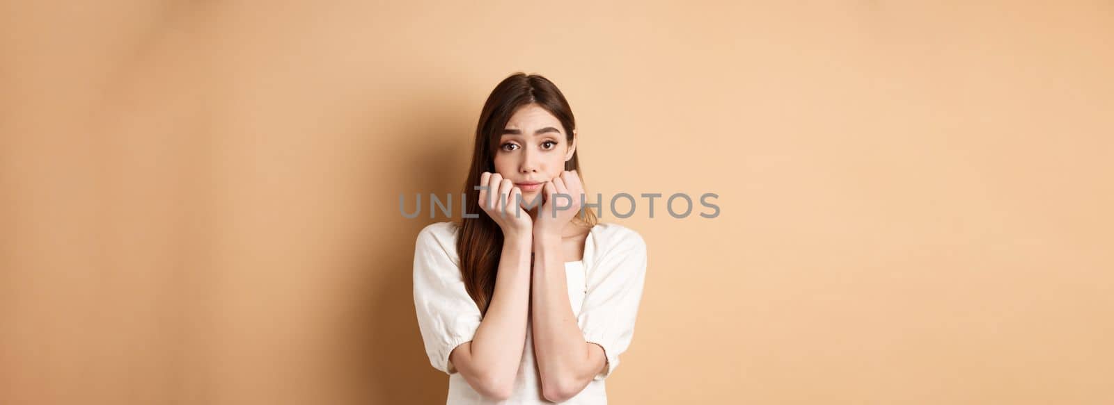 Cute and silly girl lean face on hands and looking dreamy at camera, standing romantic on beige background.