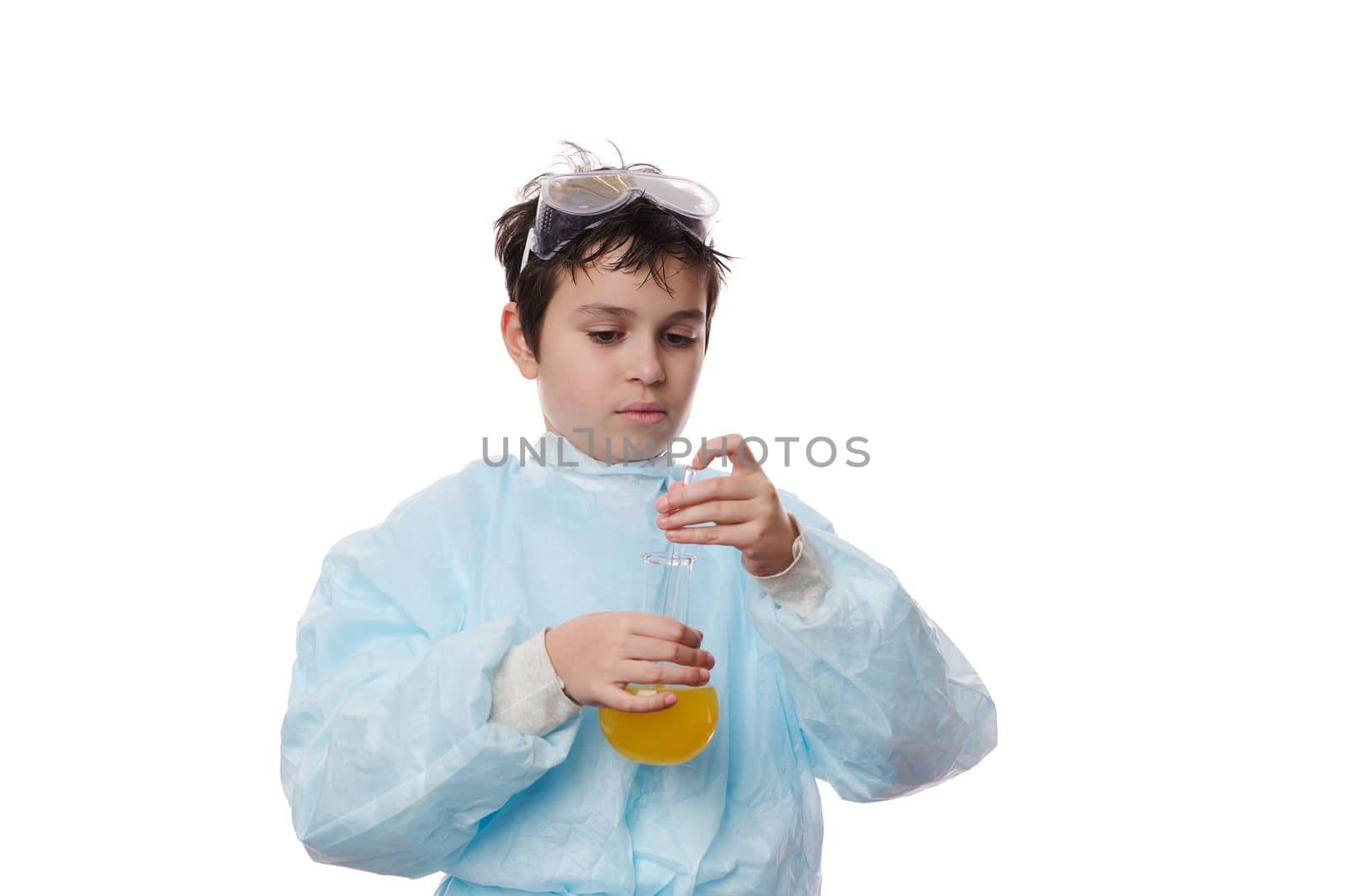 Smart teenage schoolboy in protective laboratory coat, conducts a scientific experiment in a chemistry lesson, using a glass rod, mixes reagents with chemicals in lab flask, isolated on white backdrop