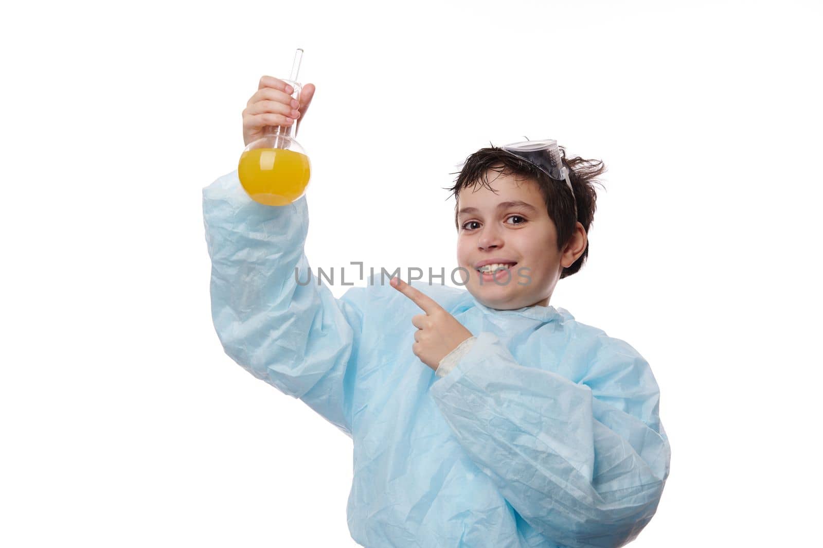 Handsome smart teenage boy in lab coat, points with index finger at a glass laboratory flask with chemical solution, conducting scientific experiments at chemistry class, isolated on white background